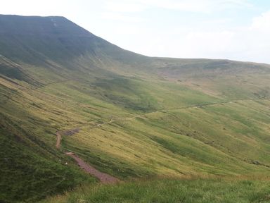 A grassy hillside with a winding dirt path leading up and across the slopes at Ty Twmp Tump Cottage in Tal-Y-Bont-On-Usk