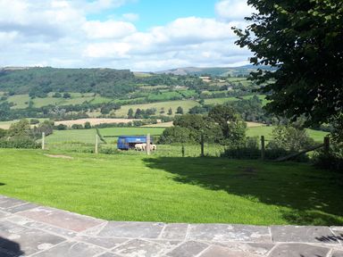 A grassy backyard with a stone patio and a fenced area containing a blue shelter and sheep with hills in the background at Ty Twmp Tump Cottage in Tal Y Bont On Usk