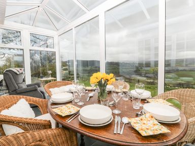 A dining table set with plates glasses and cutlery in a conservatory with wicker chairs and a view of the countryside at Ty Twmp Tump Cottage in Tal Y Bont On Usk