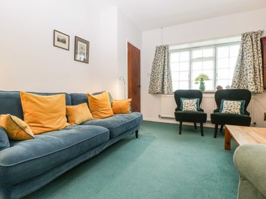 A sitting room with a blue sofa with orange cushions two black chairs by a window with patterned curtains at Ty Twmp Tump Cottage in Tal Y Bont On Usk