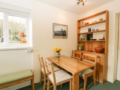 A dining area with a wooden table, four chairs, a wooden shelving unit with pottery, a bench under a window with a fruit basket on the sill at Ty Twmp Tump Cottage in Tal-Y-Bont-On-Usk