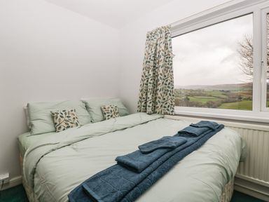 A bedroom with a made bed with green bedding and patterned cushions beside a large window showing a countryside view at Ty Twmp Tump Cottage in Tal-Y-Bont-On-Usk