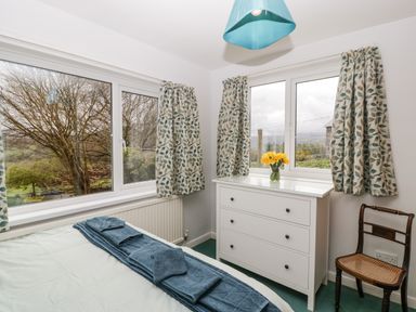 A bedroom with a bed, blue towels, a white chest of drawers with flowers, a wooden chair, and windows with curtains at Ty Twmp Tump Cottage in Tal Y Bont On Usk