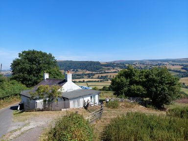 A white cottage with chimneys near a tree and a road with hills in the background at Ty Twmp Tump Cottage in Tal-Y-Bont-On-Usk