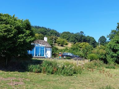A white cottage with a thatched roof and a blue extension next to a parked black car in a grassy area with trees and hills in the background at Ty Twmp Tump Cottage in Tal-Y-Bont-On-Usk