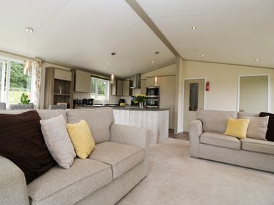 A living room with beige sofas and yellow pillows next to an open kitchen with a kitchen island at Hill View Lilac Lodge in Wareham