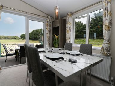 A dining area with a marble table set for six and gray chairs by windows overlooking a green outdoor area at Hill View Lilac Lodge in Wareham