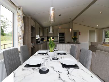 A dining table set with plates and glasses next to a kitchen with cabinets and appliances at Hill View Lilac Lodge in Wareham