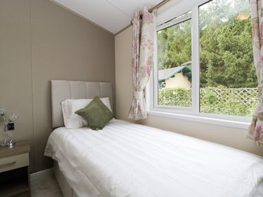 A bedroom with a single bed white bedding green cushion bedside table lamp and window with floral curtains at Hill View Lilac Lodge in Wareham