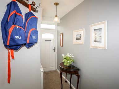 A hallway with a white front door two blue backpacks hanging on wall hooks a small wooden table with a vase of white flowers and two framed pictures on a gray wall at Silva in Penmon