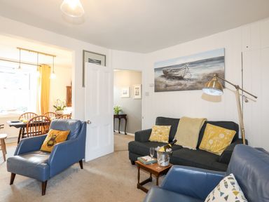 A living room with blue armchairs a dark sofa yellow cushions and a painting of a boat with a dining area visible at Silva in Penmon