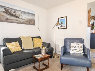 A living room with a grey sofa and yellow cushions a blue armchair a wooden side table with books and a champagne bucket a floor lamp and paintings on white walls at Silva in Penmon