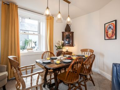 A dining room with a wooden table set with plates and glasses of juice wooden chairs yellow curtains and a small cabinet on the wall with a pitcher at Silva in Penmon