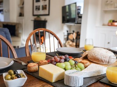 A dining table with cheese, grapes, bread, olives, and glasses of orange juice in a room with wooden chairs and a fireplace at Silva in Penmon