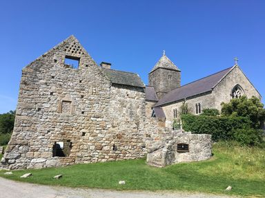 A stone building with a tower and sloped roofs surrounded by grass and bushes at Silva in Penmon