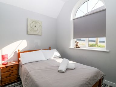A bedroom with a bed covered by a gray duvet two white pillows a wooden bedside table with a lamp and a window with a gray blind and a stuffed animal on the sill at Seagull Cottage in Bude