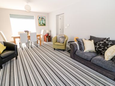 A living room with a striped carpet a dining table with chairs a green armchair a grey sofa with cushions and a painting on the wall at Seagull Cottage in Bude