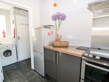 A kitchen with a refrigerator, washing machine, stove, and countertop with flowers and basket at Seagull Cottage in Bude