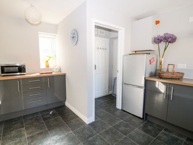 A kitchen with grey cabinets a microwave a refrigerator flowers in a vase and a wicker basket at Seagull Cottage in Bude