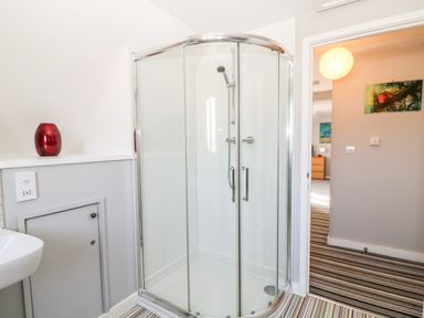 A bathroom with a glass shower enclosure a red vase on a ledge and a view into a striped carpeted room at Seagull Cottage in Bude