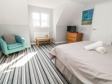 A bedroom with a bed with towels on it a turquoise armchair a wooden table with flowers and a dresser with a TV at Seagull Cottage in Bude