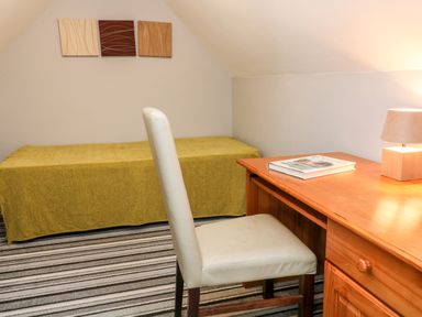 A bedroom with a single bed covered with a yellow blanket a wooden desk with a book and lamp and a white upholstered chair at Seagull Cottage in Bude