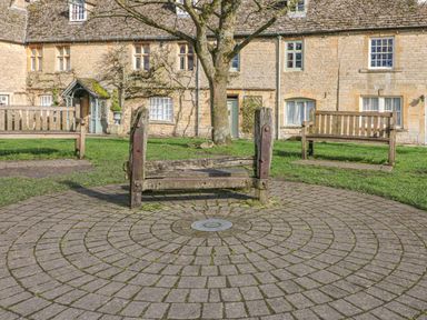 A circular stone paved area with wooden benches and a tree in front of stone cottages at Coln Cottage in Stow On The Wold