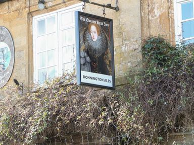 A pub sign with a portrait of a queen and text the queens head inn donnington ales on a stone building with windows and climbing plants