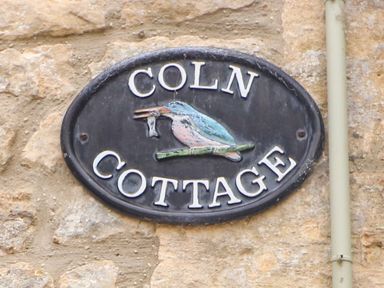 A wall-mounted oval plaque with a bird holding a fish and text reading Coln Cottage on a stone wall at Coln Cottage in Stow-On-The-Wold