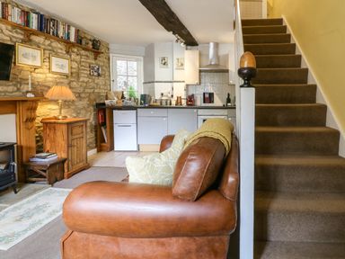 A living area with a brown leather sofa next to carpeted stairs and a kitchen with light gray cabinets at Coln Cottage in Stow-On-The-Wold