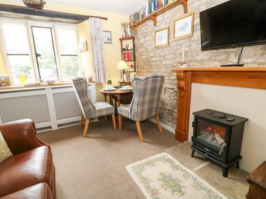 A sitting area with two checkered chairs at a small round table near a window and an electric stove under a mounted television at Coln Cottage in Stow On The Wold