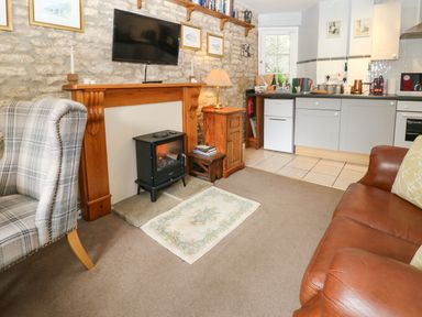 A living room with a leather sofa a plaid armchair a fireplace with a stove television kitchen cabinets and appliances at Coln Cottage in Stow-On-The-Wold