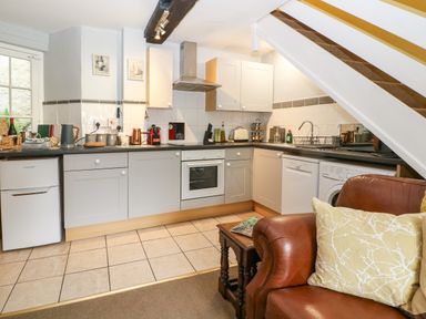 A kitchen with white cabinets and appliances including oven dishwasher and washing machine with a brown chair and cushion in the foreground at Coln Cottage in Stow-On-The-Wold