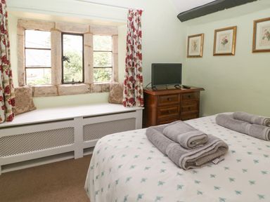 A bedroom with a bed folded towels a wooden dresser with a television and a window seat with cushions at Coln Cottage in Stow-On-The-Wold