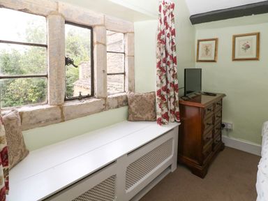 A bedroom corner with a window seat cushions a wooden chest of drawers with a television and floral curtains at Coln Cottage Stow-On-The-Wold