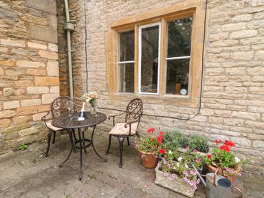 An outdoor seating area with a metal table and two chairs next to a stone wall with a window and several flower pots at Coln Cottage in Stow-On-The-Wold
