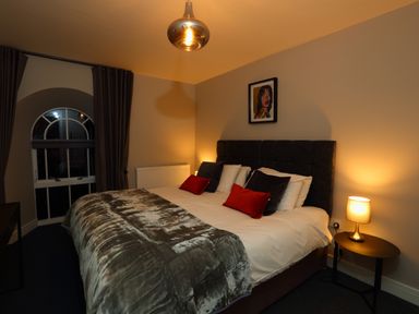 A bedroom with a double bed with pillows and cushions a bedside table with a lamp and an arched window at The Poppy Fields Abode in Dyserth