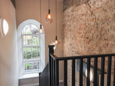 A stairway with black railing next to a stone wall and arched window with hanging pendant lights at The Sigma Penthouse in Dyserth