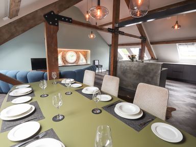 A dining table set with plates and glasses in an open plan room with exposed beams and a kitchen in the background at The Sigma Penthouse in Dyserth