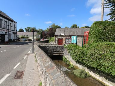 A street with a stone wall alongside a narrow stream and buildings including a pub and a red phone booth at The Sigma Penthouse in Dyserth