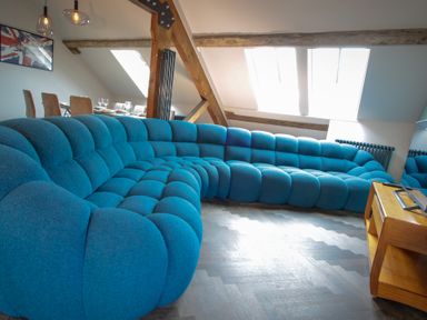 A living room with a large blue sectional sofa wooden beams and a dining table with chairs at The Sigma Penthouse in Dyserth