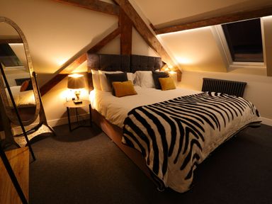 A bedroom with a large bed covered with zebra patterned blanket, bedside tables with lamps, a standing mirror, and wooden beams on the ceiling at The Sigma Penthouse in Dyserth