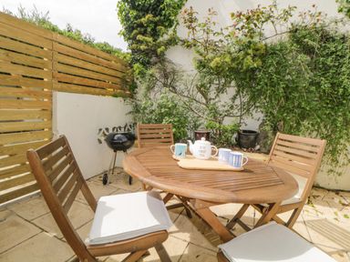 A patio with a wooden table and chairs with cushions a teapot and mugs on the table and a small barbecue grill next to a wooden fence at Saltwater Cottage in Babbacombe