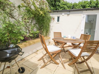 A patio with a round wooden table and four wooden chairs with cushions next to a charcoal grill and plants at Saltwater Cottage in Babbacombe