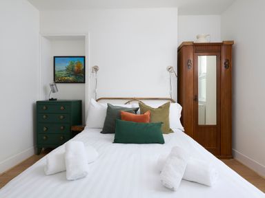 A bedroom with a made bed with five pillows and two rolled towels a green chest of drawers with a lamp and a painting and a wooden wardrobe with a mirror at Saltwater Cottage in Babbacombe