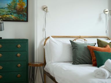 A bedroom with a bed with white sheets and multiple colored pillows next to a green chest of drawers and a small wooden side table at Saltwater Cottage in Babbacombe