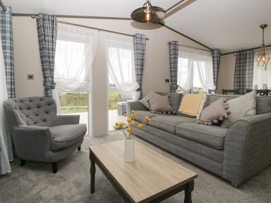 A living room with a grey sofa and armchair a wooden coffee table with flowers and plaid curtains at 14 Fairways in Burnham-On-Sea