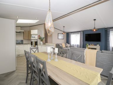 A dining area with a wooden table and six chairs next to a kitchen and living room with grey sofas and a wall-mounted TV at 14 Fairways in Burnham-On-Sea