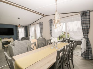A dining area with a wooden table and chairs next to a living room with grey sofas and a wall-mounted television at 14 Fairways in Burnham-On-Sea
