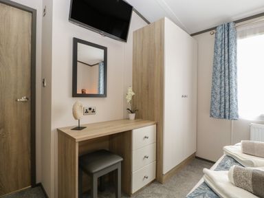 A bedroom with a wooden desk, gray stool, wall mirror, wardrobe, TV, blue patterned curtains, and folded towels on a bed at 14 Fairways in Burnham-On-Sea